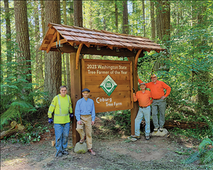 Four people stand in the forest alongside a large wooden sign that says 2023 Washington State Tree Farmer of the Year Coburg Farm